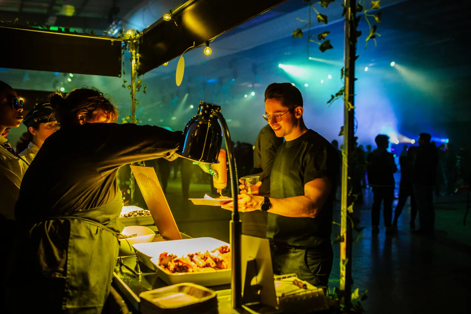 Attendees at a vibrant evening event interact with a food buffet setup under stylish lighting. The scene features a person serving appetizers while others gather around, creating an engaging social atmosphere.