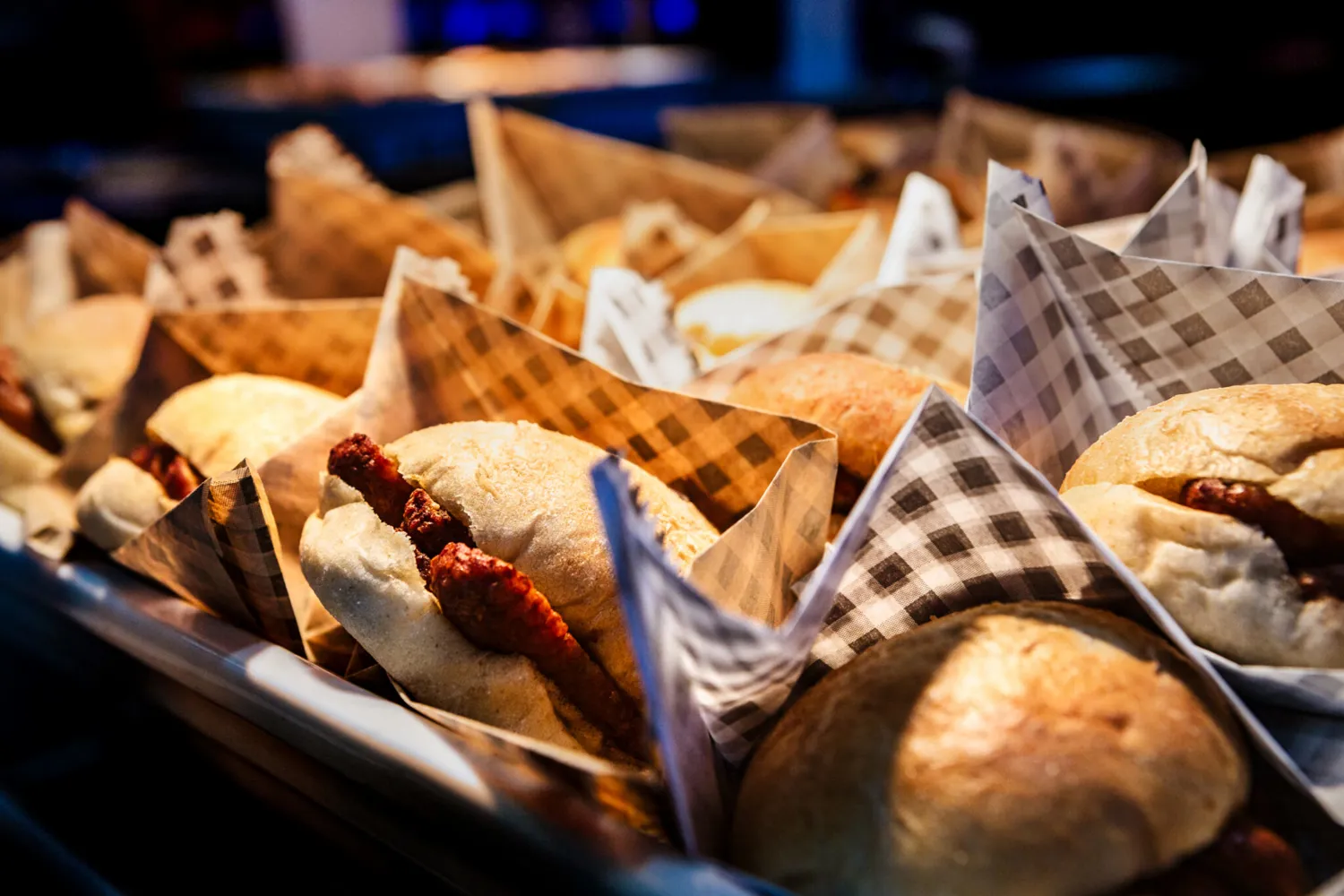 Delicate baked goods wrapped in checkered paper napkins, featuring small toppings, arranged in a tray. The presentation highlights a variety of pastries ready for serving at an event.